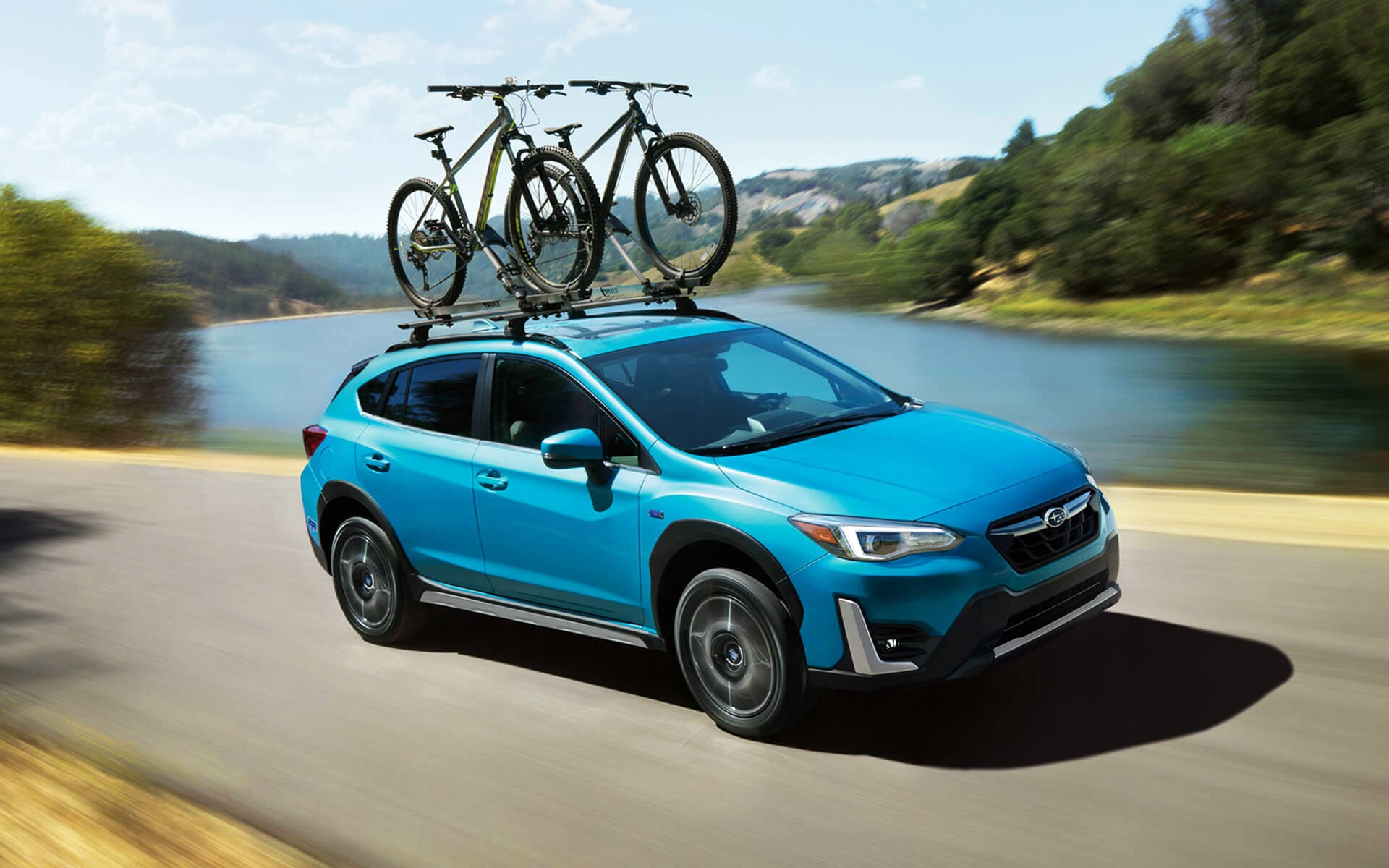 A blue Crosstrek Hybrid with two bicycles on its roof rack driving beside a river | Tindol Subaru in Gastonia NC